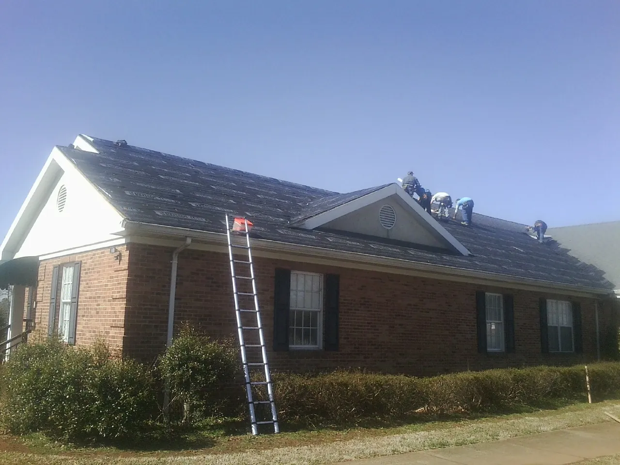 Crew tearing shingles off a brick ranch roof with a ladder set — Metal Roof Repair work by our crew in Holden